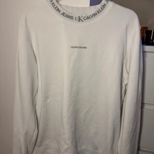 Vit sweatshirt från Calvin Klein Jeans - Vit sweatshirt från Calvin Klein Jeans med logga tryckt på bröstet och runt halsringningen. Tröjan har en klassisk passform, långa ärmar och är tillverkad i mjuk bomull. Perfekt för en clean och stilren look.
