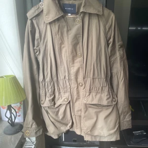 Beige parkajacka från 365 Sunshine - Snygg beige parkajacka från 365 Sunshine med stora fickor, dragkedja och knappar framtill. Jackan har markerad midja, klassisk krage och detaljer på axlarna. Perfekt för dig som gillar en stilren och avslappnad look.