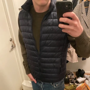 Marinblå dunväst från Uniqlo - Säljer en svart dunväst från Uniqlo i storlek M. Västen är quiltad, har dragkedja framtill och är lättviktig. Perfekt att slänga över en hoodie eller tröja när det är kyligt ute. Klassisk och stilren design som funkar till det mesta.