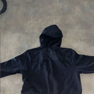 Svart hoodiejacka från Under Armour - Svart hoodiejacka från Under Armour med dragkedja framtill och blå logga på bröstet. Jackan har huva, långa ärmar och är tillverkad i mjukt syntetmaterial. Perfekt för träning eller chill. Snygg och enkel design med praktiska fickor.