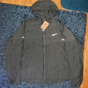 Grå Nike Miller vindjacka - Svart vindjacka från Nike med huva och dragkedja framtill. Jackan har två sidofickor och reflekterande detaljer på ärmarna samt klassisk Nike-logga på bröstet. Lätt och smidig modell i syntetmaterial, perfekt för blåsiga dagar.