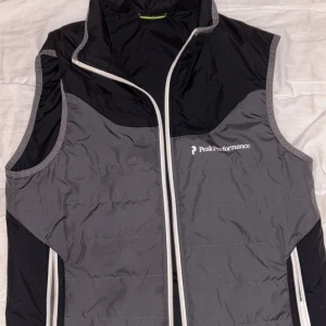 Grå och svart dunväst Peak Performance - Snygg dunväst från Peak Performance i grått och svart med vit dragkedja och logga på bröstet. Västen har hög krage och är lätt vadderad, perfekt för lager-på-lager. Sportig look och passar till många outfits. Pris kan diskuteras 
