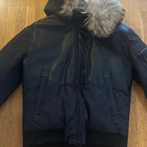 Svart bomberjacka Rockandblue med fuskpäls - Snygg svart bomberjacka från Rockandblue med fuskpäls på huvan. Jackan har blandning av skinn och syntetmaterial, dragkedja framtill, fickor och en extra ficka på ärmen. Perfekt för kalla dagar med sin fodrade insida och coola look.