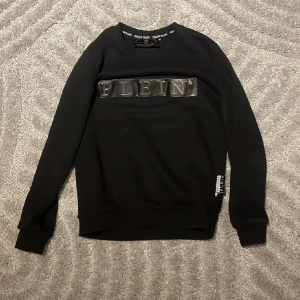 Svart sweatshirt från Philipp Plein - Svart sweatshirt från Philipp Plein med metallic-logga framtill och ribbade muddar. Klassisk rund halsringning och diskret logodetalj bak i nacken. Snygg och stilren tröja som sticker ut med sin tydliga branding.