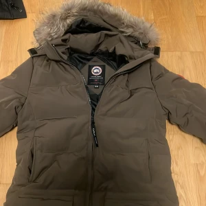 Brun dunjacka från Canada Goose - Säljer en brun dunjacka från Canada Goose med huva och snygg fuskpäls. Jackan har dragkedja framtill, klassisk logga på ärmen och är perfekt för kalla vinterdagar. Pris kan diskuteras 