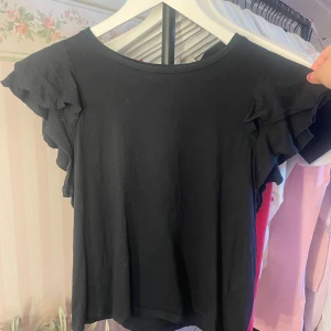 Svart volangtopp från Amisu - Svart topp från Amisu i mjuk bomull med volangdetaljer på ärmarna. Klassisk rund halsringning och korta ärmar med dubbla volanger som ger en söt och trendig look. Perfekt att styla med jeans eller kjol för en enkel men snygg outfit.