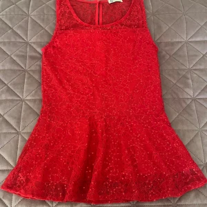 Röd spetstopp med volang från Cubus - Supersöt röd topp i spets från Cubus, med ärmlös design och volang nertill. Toppen har blommigt spetsmönster och små paljetter som ger extra glans. Perfekt för dig som vill sticka ut med en färgstark och feminin topp.