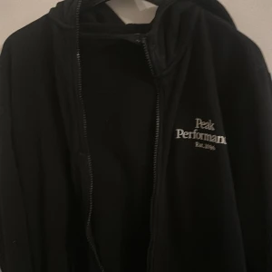 Svart hoodie från Peak Performance - Svart hoodie från Peak Performance med vit broderad logga på bröstet. Tröjan har dragkedja framtill, huva och långa ärmar. Perfekt för dig som gillar en stilren och sportig look.