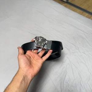 Svart Versace bälte med Medusa-spänne - Säljer ett svart bälte från Versace med ikoniskt Medusa-huvud i silver som spänne. Bältet är gjort i slätt skinn och har en klassisk, stilren design som lyfter vilken outfit som helst. Perfekt accessoar för dig som gillar lyxiga detaljer och statement pieces.