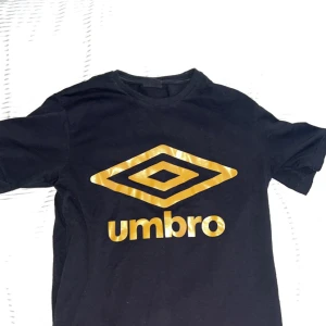 Svart t-shirt med guldtryck från Umbro - Svart t-shirt från Umbro med stort guldigt logotryck på bröstet. Klassisk rund hals och korta ärmar. Skön passform och tillverkad i mjuk bomull. Perfekt för dig som gillar sportiga och stilrena plagg med en touch av street, Har aldrig använt. 