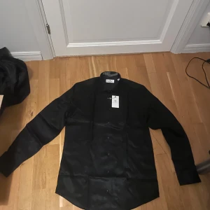 Svart skjorta från Tee Jays - Stilren svart skjorta från Tee Jays med klassisk krage och lång ärm. Skjortan har knappar framtill och är tillverkad i ett mjukt bomullsmaterial som känns skönt mot huden. Perfekt för dig som gillar en clean och enkel look. Har i fler färger också 