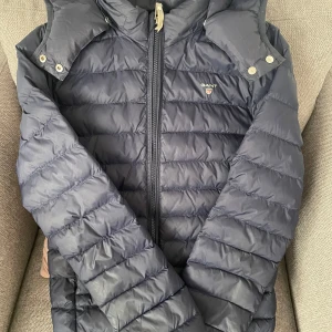 Mörkblå pufferjacka från Gant - Snygg mörkblå pufferjacka från Gant med huva och dragkedja framtill. Jackan har quiltad design och Gant-logga på bröstet. Perfekt för kyliga dagar och riktigt skön att bära. Klassisk look som funkar till det mesta. Bara att kontakta för fler bilder 
