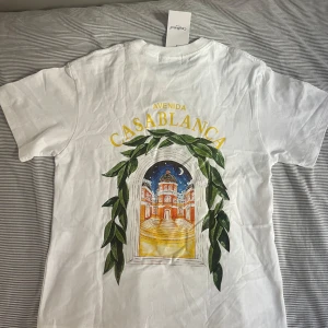 Vit t-shirt från Casablanca Tennis Club - Vit t-shirt från Casablanca Tennis Club med färgglad grafisk print av en byggnad och gröna blad på både fram- och baksidan. Klassisk passform och rund halsringning. Tillverkad i mjuk bomull, perfekt för en clean och stilren look.