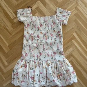 Vit blommig kortklänning med volang - Supersöt kortklänning i vit bas med blommigt mönster i rosa, grönt och gult. Klänningen har smockad överdel, korta ärmar och en volangig kjol. 