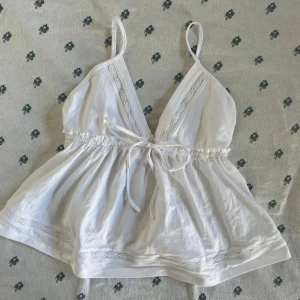 Vit babydoll linnetopp med spets från Gina Tricot - Supersöt vit babydoll linnetopp från Gina Tricot med smala axelband, v-ringning och spetsdetaljer längs urringningen och nederkanten. Toppen har knyt i midjan och en lätt volangig, luftig passform. Perfekt för varma dagar och en fräsch look.