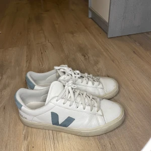 Vita Veja sneakers med blå detalj - Snygga vita Veja sneakers i skinn med blå V-logga på sidan och blå detalj vid hälen. Klassisk låg modell med snörning och platt sula. Perfekta för en clean och stilren look, passar till det mesta i garderoben.