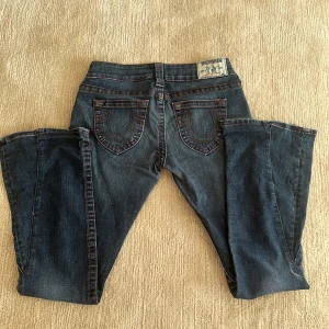 Vintage True Religion bootcut jeans blå, stl 27 - Säljer ett par vintage blå jeans från True Religion med bootcut passform. Jeansen har röda kontrastsömmar, ikoniska bakfickor med broderi och patch på linningen. Tillverkade i USA i mjukt denimtyg. Köpta på second hand och säljer då de är lite för korta för mig (jag är 171)