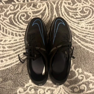 Nike Phantom GT fotbollsskor svart/blå - Nästan helt nya Säljer ett par Nike Phantom GT fotbollsskor i svart med blå detaljer och grå sula. Skorna har snörning och är designade för inomhusspel med slitstark syntetisk ovandel och greppvänlig yttersula. Snygg och modern look med tydliga Nike-loggor på sidorna.