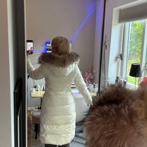 Vit lång dunjacka med pälskrage Moncler - Säljer en lång, vit dunjacka från Moncler med stor fluffig pälskrage i brunt. Jackan har dragkedja framtill, två fickor med dragkedja och huva. Perfekt för kalla dagar och riktigt snygg siluett.