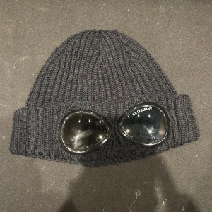 Svart goggle-mössa från C.P. Company - Svart ribbstickad mössa från C.P. Company med ikoniska goggle-linser framtill. Mössan är tillverkad i ett tjockt, stickat material som ger en cool och urban look. Perfekt accessoar för dig som vill sticka ut och addera streetstyle till outfiten. Pris går att diskuteras