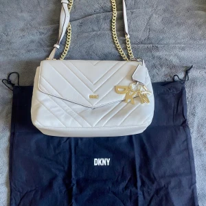 Vit quiltad axelväska från DKNY - Snygg vit axelväska från DKNY i quiltat skinn med guldfärgade kedjedetaljer och justerbar axelrem. Väskan har dekorativa DKNY-bokstäver i metall och klassisk kuvertformad lock. Perfekt för dig som vill ha en stilren och trendig accessoar.