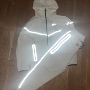 Nike Tech Fleece dress med reflexdetaljer - Nike Tech Fleece dress i färgen vit med svarta dragkedjor och feta reflexdetaljer på bröstet och ärmarna. Tröjan har huva, hel dragkedja och två fickor framtill. Perfekt för dig som vill sticka ut och hålla stilen när det är mörkt ute.