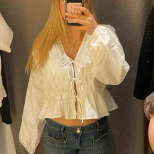 Vit blus med knyt från Gina  - Supersöt vit blus från GinaTricot med tre knyt framtill och volangkant nedtill. Blusen har lång ärm, v-ringning och är lätt croppad. Perfekt för dig som gillar romantisk och trendig stil. Tunn och luftig.