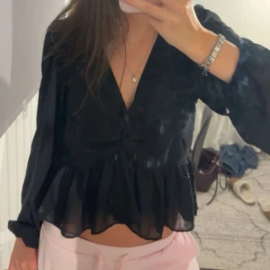 Svart blus med volang och v-ringning - Säljer en svart blus med v-ringning och volang nertill. Blusen har långa ärmar och är tillverkad i ett tunt, lätt och lite transparent material som ger en luftig känsla. Perfekt att styla med jeans eller kjol för en snygg look.