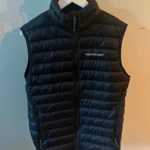Svart dunväst Peak Performance - Svart dunväst från Peak Performance med quiltad design och hög krage. Västen har dragkedja framtill och två sidofickor med dragkedja. Perfekt för lager-på-lager och har en stilren look med diskret logga på bröstet.