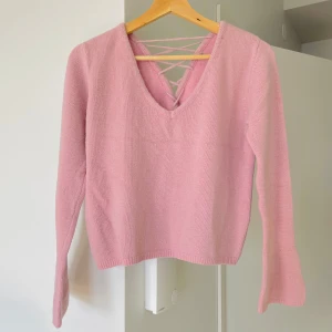 Rosa stickad tröja med snörning - En stickad tröja med ull, mjuk och bekväm att bära. Axelbredd: 39 cm, längd: ca 49 cm, ärmlängd: 59 cm. Storlek M, designad med korsade snörningsdetaljer som kan justeras för passform. Utmärkt skick. 