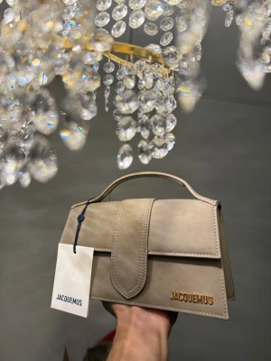 Jacquemus the large bambino - Snygg beige handväska från Jacquemus i mocka med stilren, rektangulär form och guldfärgad logga. Väskan har ett topphandtag och medföljande axelrem med gulddetaljer. Perfekt för dig som gillar design och exklusiva accessoarer.