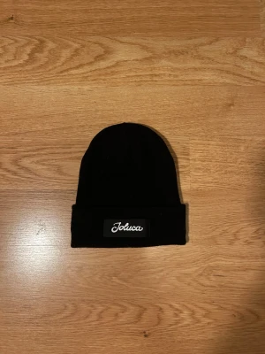 Svart mössa från Joluca - En stilren svart mössa från Joluca med uppvikt kant och en svart patch med vit broderad logga framtill. Mössan är ribbstickad och gjord i mjukt material som håller dig varm. Perfekt accessoar för kalla dagar och enkel att matcha med olika outfits. 