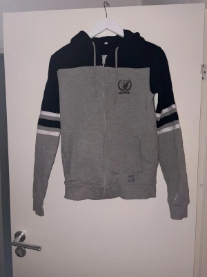 Grå och svart hoodie från FBK - Snygg zip-hoodie från FBK i grått och svart med vita och svarta ränder på ärmarna. Broderade detaljer med texten 1932 och FBK på bröstet och ärmen. Mjuk bomullsmix, två fickor fram och dragsko i huvan. Perfekt för dig som gillar sportig stil.