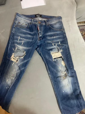 Dsquared2 blå  jeans  - Säljer ett par blå jeans från Dsquared2 med coola slitningar och stora hål på benen. Jeansen har färgstänk i rött, gult och vitt som ger en unik street vibe. Klassisk femficksmodell med knapp och dragkedja. 