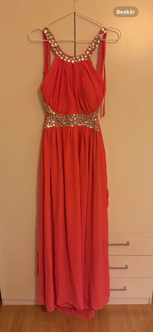 Lång korallfärgad klänning med paljettdetaljer - Snygg långklänning i korall med glittriga paljetter längs axelband och midja. Klänningen har ärmlös design och ett mjukt, fallande tyg som ger en elegant look. Perfekt för bal eller festliga tillfällen.