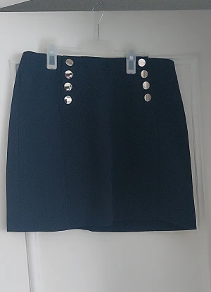 Mörkblå kortkjol med knappar från H&M - Snygg mörkblå kortkjol från H&M med dekorativa silvriga knappar framtill. Kjolen har en rak passform och är tillverkad i ett strukturerat syntetmaterial. Dragkedja bak för enkel påklädning. Perfekt för dig som gillar stilrena detaljer.