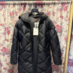 Svart Moncler dunjacka med huva - Säljer en svart dunjacka från Moncler med snyggt quiltat mönster och huva. Jackan har lång ärm, dragkedja och Moncler-logga på ärmen. Perfekt för kalla dagar och har en clean, stilren look som funkar till allt. Jackan är tillverkad i polyester och dun.