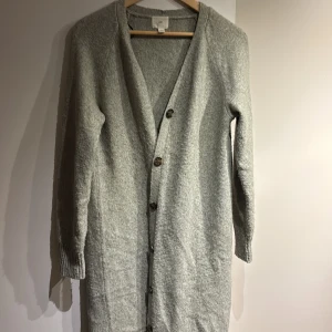 Grå lång kofta från H&M - Säljer en grå, lång och mysig kofta från H&M med v-ringning och knappar framtill. Koftan är stickad och har långa ärmar med ribbade muddar. Perfekt att slänga på sig när det är lite kyligt ute.