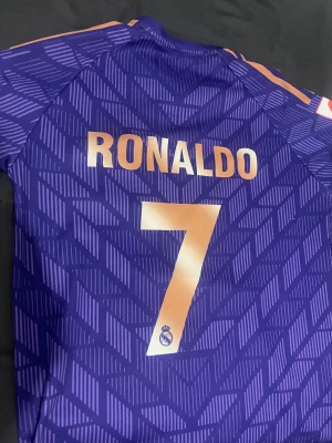 Real Madrid Ronaldo #7 Adidas t-shirt - Säljer en fet Real Madrid t-shirt från Adidas med Ronaldo och nummer 7 på ryggen. Tröjan är lila med guldiga detaljer och har ett grafiskt mönster över hela. Kortärmad med rund hals, Real Madrid-emblem och Emirates Fly Better tryck på bröstet. Perfekt för fotbollsfans!