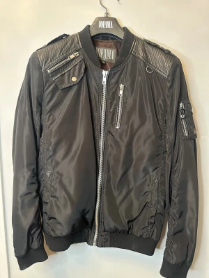 Svart bomberjacka från Jofama - Svart bomberjacka från Jofama med coola detaljer som quiltade axelpartier, flera dragkedjor och fickor. Jackan har ribbade muddar vid ärmar och midja, samt snygga metallknappar. Materialet är blankt och känns lätt, med inslag av skinn på axlarna. Har en liten skada på höger ficka. 