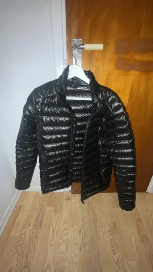 Svart dunjacka från J.Lindeberg - Svart, glansig dunjacka från J.Lindeberg med horisontella quiltade paneler och dragkedja framtill. Jackan har en stilren, smal passform och hög krage som ger en modern look. Perfekt för kalla dagar när du vill hålla stilen.