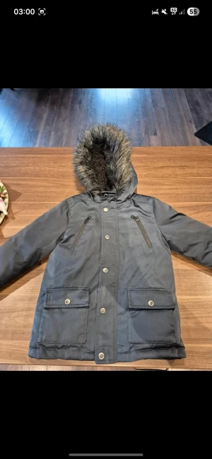 Mörkblå parkajacka med fuskpäls Minoti - Parkajacka från Minoti i mörkblått med blått quiltat foder och huva med fuskpäls. Jackan har två stora fickor med knappar, två bröstfickor med dragkedja och mysigt teddyfoder i huvan. Perfekt för kalla dagar.