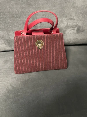 Randig röd handväska med gulddetaljer - Snygg handväska från Ted Baker i mörkröd och vinröd ton med stilrent randigt mönster. Väskan har guldfärgad låsdetalj, två handtag och insida med extra fack. Materialmix av skinn och textil ger en lyxig känsla. Perfekt för dig som vill sticka ut med en unik accessoar.