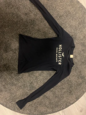 Svart långärmad tröja från Hollister - Tunn snygg svart långärmad tröja från Hollister med vit logotyp och text på bröstet. Perfekt för dig som gillar enkel och stilren design. Välanvänd men i fint skick. Lappen på insidan bortklippt. Hör av dig vid frågor💕