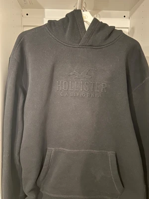 Svart hoodie från Hollister - Svart hoodie från Hollister med broderad logga och text på bröstet. Tröjan har huva, magficka och långa ärmar. Mjuk och skön bomullskänsla, perfekt för chill dagar. Enkel och stilren design som funkar till jeans eller mjukisbyxor. Tröjan har en sjukt bra pass form, dock ett litet hål vid ena ärmen. Detta är den enda defekten, vilken inte syns. Om man vill kan man lätt sy med ett par stygn bara!