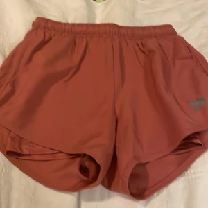 Rostfärgade träningsshorts 157 X-Small - Säljer ett par snygga rostfärgade träningsshorts från 157 i storlek X-Small. De har elastisk midja med snörning och är tillverkade i ett lätt och snabbtorkande syntetmaterial. Perfekta för löpning eller gymmet, med sportig passform och diskret logga på benet.