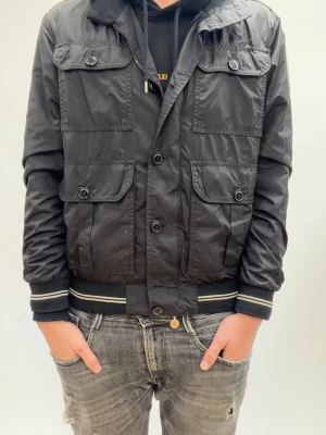 Svart vindjacka från Moncler - Svart vindjacka från Moncler med fyra stora fickor framtill och knappar. Jackan har ribbade muddar med vita ränder. Riktig shysst nu till Vintern/Höst. 