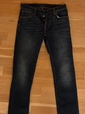 Mörkblå Nudie Jeans Skinny W29 L30 - Snygga mörkblå jeans från Nudie Jeans med skinny passform och klassiska orangea sömmar. Modellen har fem fickor, knappgylf och coola detaljer på bakfickorna. Perfekt för dig som gillar en smal siluett och stilren look. Tveka inte av att höra av er! Priset kan diskuteras vid snabb affär!