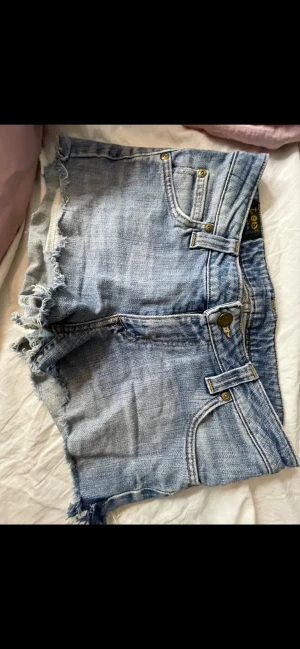 Blå jeansshorts från Lee med rå kant - Säljer ett par blå jeansshorts från Lee med klassisk femficksdesign och rå, fransig kant nedtill. Shortsen har låg midja, slitna detaljer och Lee-logga på bakfickan och linningen. 