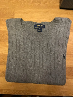 Grå stickad tröja Polo Ralph Lauren XL - Snygg grå stickad tröja från Polo Ralph Lauren i storlek XL. Tröjan har klassiskt kabelstickat mönster, rund hals och den ikoniska broderade loggan på bröstet. Perfekt för dig som gillar stilrena och tidlösa plagg. Liten fläk på tröjan